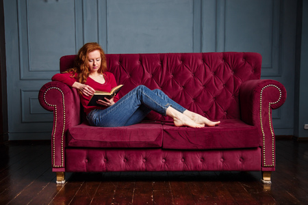 Rich young redhead woman reading a book on red velvet sofa.の写真素材