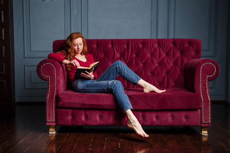 Rich young redhead woman reading a book on red velvet sofa.の写真素材