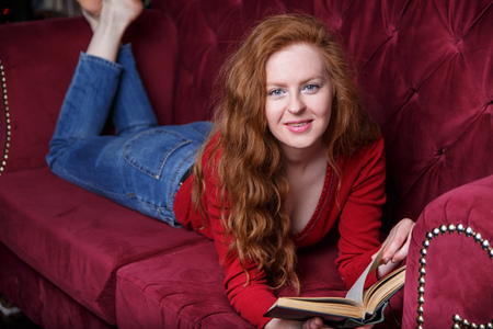 Rich young redhead woman reading a book on red velvet sofaの写真素材