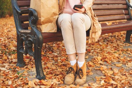 Young lady sitting on bench with paper bag and phone in autumn park.の写真素材