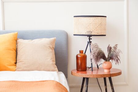 Stylish lamp on wooden nightstand next to flower in big glass vase in scandinavian bedroom interior.の写真素材