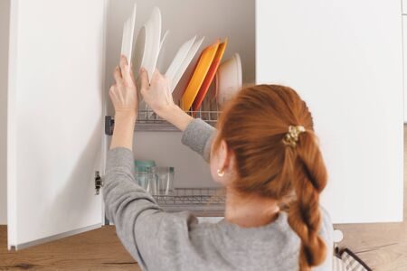 Opening kitchen cabinet door, woman putting plates into itの写真素材