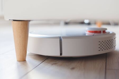 Robot vacuum cleaner is vacuuming under the bed. Cleaning in inaccessible places. Automation and smart home, modern technologyの写真素材