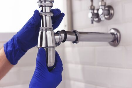 Plumber repairs and maintains chrome siphon under the washbasin. Plumber at work in bathroom, plumbing assemble and install concept.の写真素材