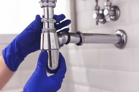 Plumber repairs and maintains chrome siphon under the washbasin. Plumber at work in bathroom, plumbing assemble and install concept.の写真素材