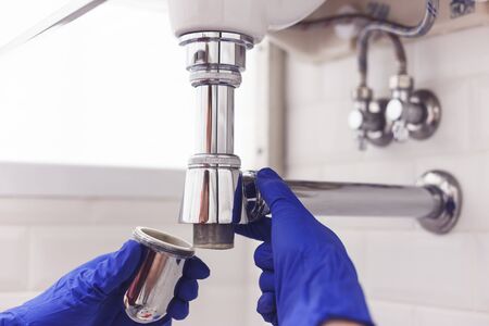 Plumber repairs and maintains chrome siphon under the washbasin. Plumber at work in bathroom, plumbing assemble and install concept.の写真素材