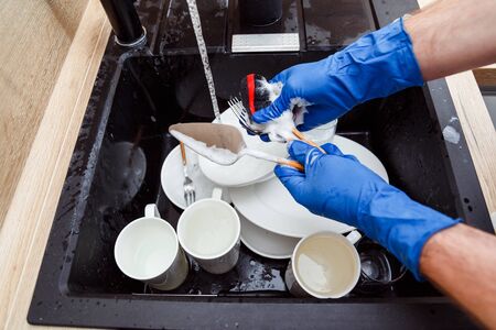 Washing of the dishes - man hands in gloves washing with sponge and rinsing dishes under running water in the sinkの写真素材