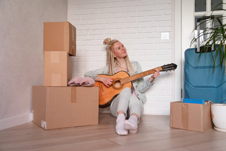 An attractive young woman is sitting on a couch and playing the guitar. Moving boxes can be seen in...