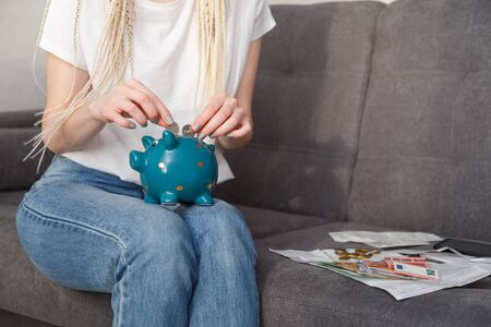 Young woman putting coins into money box sitting on sofa at homeの写真素材
