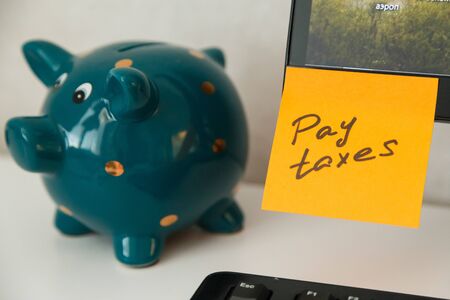 Reminder pay taxes sticked on monitor at freelancers workplace. Finance planning concept. Money-box for savings on backgroundの写真素材