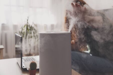 Woman freelancer uses a household humidifier in the workplace to maintain relative humidity and microclimate in the workplace of the home office with a laptop and documents.の写真素材