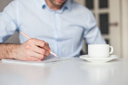 Man writes to-do list in notebook, working at home. Drink coffee, keep a pencil. Wearing blue casual shirt. No gadgets.の写真素材