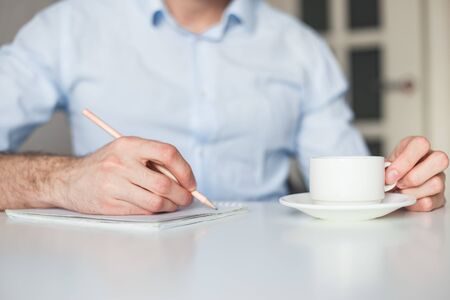 Man writes to-do list in notebook, working at home. Drink coffee, keep a pencil. Wearing blue casual shirt. No gadgets.の写真素材