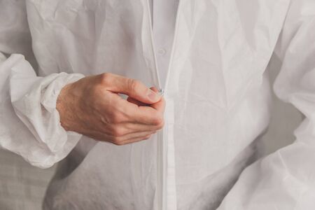 Doctor epidemiologist puts on white protective suit, fastens a zipper, hands close-up. Hands in protective rubber gloves. Personal protective equipment concept.の写真素材