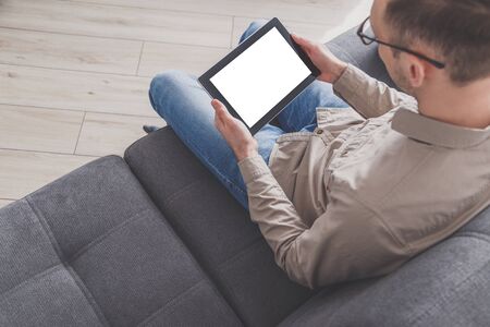 Internet surfing and conference calling. A man uses a tablet computer while sitting on a sofa at home. Dressed in everyday clothes. Tablet with blank screen.の写真素材