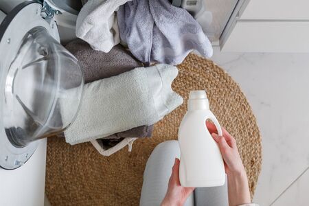 Woman holds bottle with liquid laundry detergent before washing clothes. Washer and clothes ready for laundry. View from above.の写真素材