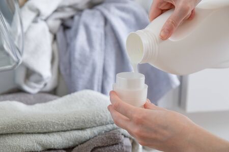 Close up of female hands pouring liquid laundry detergent into cap. Washer machine and clothes with wicker basket in background.の写真素材