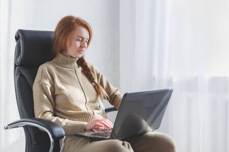Young caucasian woman sitting in leather armchair using laptop working from home, freelance, social distancing and remote work concept.の写真素材