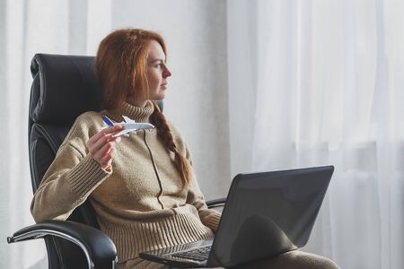 Woman dreaming about travel while self isolation at home. Caucasian girl with toy airplane and laptop sitting in armchair.の写真素材