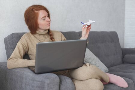 Woman dreaming about travel while self isolation at home. Caucasian girl with toy airplane and laptop sitting in armchair.の写真素材