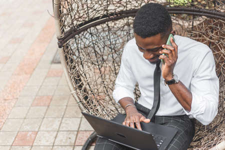 Cheerful young African man in formal wear talking on the mobile phone and working on laptop while sitting on egg chair at outdoors cafeの写真素材