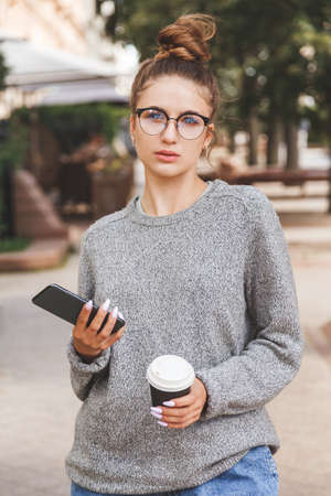 Young girl dressed in casual clothes with smartphone and a cup of takeaway coffee. Daily life of youth. People onlineの写真素材