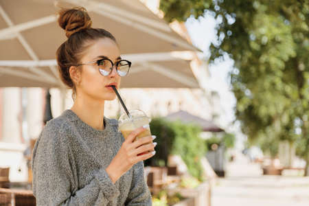 Alone young woman in eyeglasses drinking milkshake and looking away in cozy cafe outdoorの写真素材