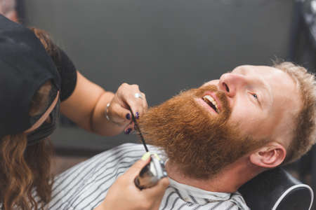 A man at a barbershop. Woman barber clipping beard and mustache. Barber woman in mask.の写真素材