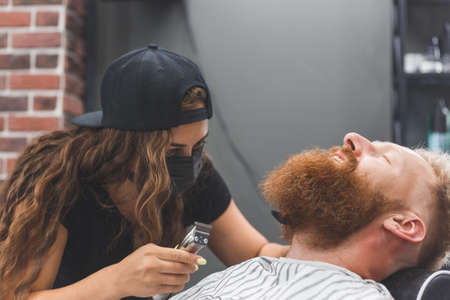 A man at a barbershop. Woman barber clipping beard and mustache. Barber woman in mask.の写真素材