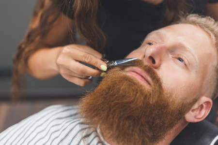 A man at a barbershop. Woman barber clipping mustache. Barber woman in mask.の写真素材