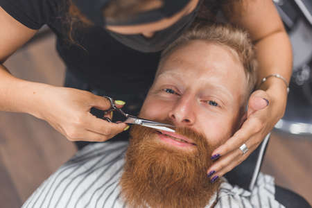 A man at a barbershop. Woman barber clipping mustache. Barber woman in mask.の写真素材