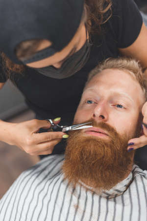 A man at a barbershop. Woman barber clipping mustache. Barber woman in mask.の写真素材