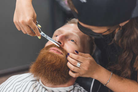 A man at a barbershop. Woman barber clipping mustache. Barber woman in mask.の写真素材