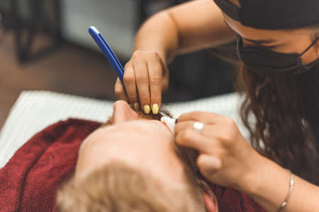 Barber woman shaves beard with a straight razor. Hairdresser equipment. Selective focus. Master in face maskの写真素材