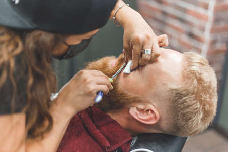 Barber woman shaves beard with a straight razor. Hairdresser equipment. Selective focus. Master in face maskの写真素材
