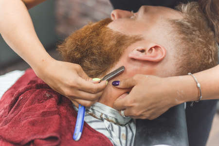Barber woman shaves beard with a straight razor. Hairdresser equipment. Selective focus. Master in face maskの写真素材