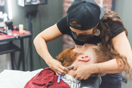 Barber woman shaves beard with a straight razor. Hairdresser equipment. Selective focus. Master in face maskの写真素材