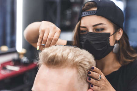 Women barber makes flat top haircut with scissors a bearded man. Service in a mask from virus.の写真素材