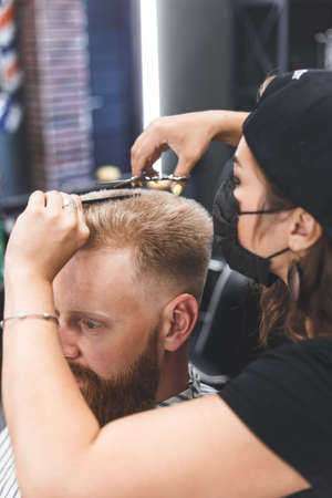 Women barber makes flat top haircut with scissors a bearded man. Service in a mask from virus.の写真素材