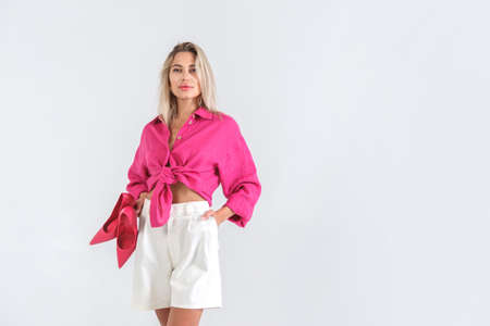 Beautiful blond woman in white shorts and pink blouse posing barefoot holding heels in studio on white background. Fashion summer lookの写真素材