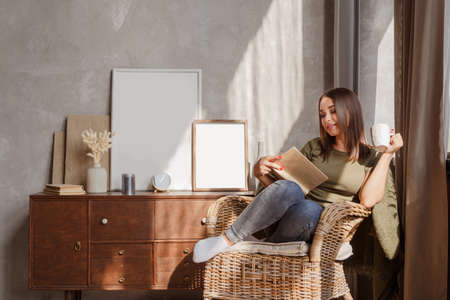 Young woman reading a book and holding cup of tea or coffee sit in rattan wicker armchairの写真素材