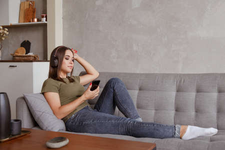 Young brunette woman enjoying the music in wireless headphones connected to smartphone, lying on couch at home aloneの写真素材