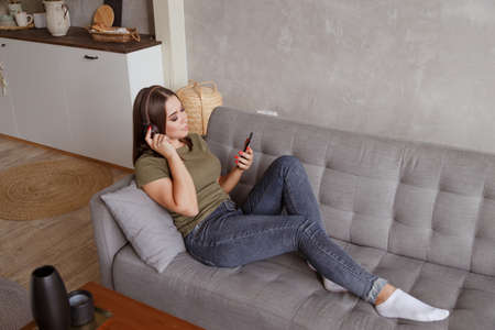 Young brunette woman enjoying the music in wireless headphones connected to smartphone, lying on couch at home aloneの写真素材