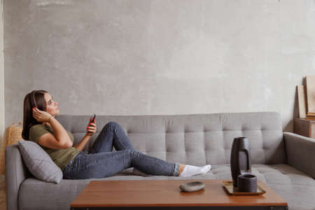 Young brunette woman enjoying the music in wireless headphones connected to smartphone, lying on couch at home aloneの写真素材