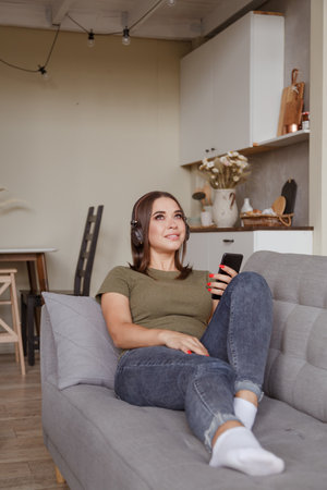 Young brunette woman enjoying the music in wireless headphones connected to smartphone, lying on couch at home aloneの写真素材