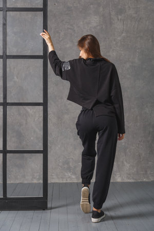 Young woman in black sportswear, pants and sweatshirt. Back view. Concept of fashionable sport outfit, indoors photo.の写真素材