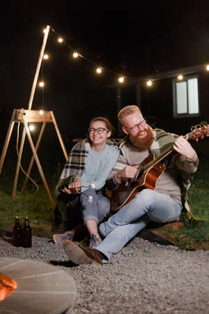Young couple, caucasian woman and red haired bearded man, on a night picnic with a guitar and marshmello have a good timeの写真素材