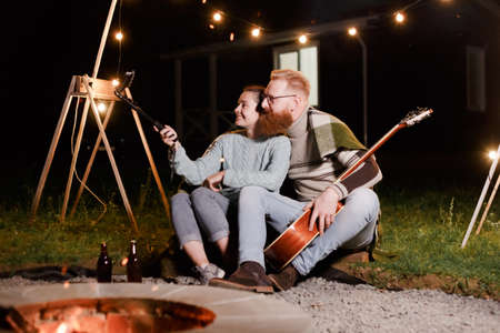 Happy caucasian couple, brunette woman and bearded man with guitar at picnic in countryside. Funny couple making selfie, smiling and have fun together.の写真素材