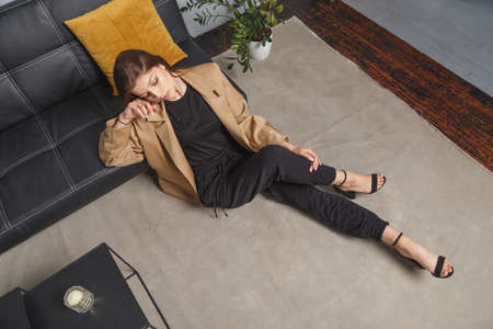 Stylishly dressed young caucasian woman in beige blazer and black pants sitting on the floor at sofa. Interior fashion portraitの写真素材