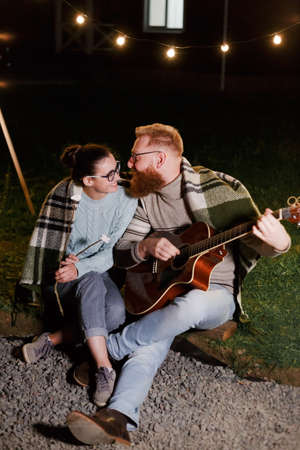 Young couple, caucasian woman and red haired bearded man, on a night picnic with a guitar and marshmello have a good timeの写真素材